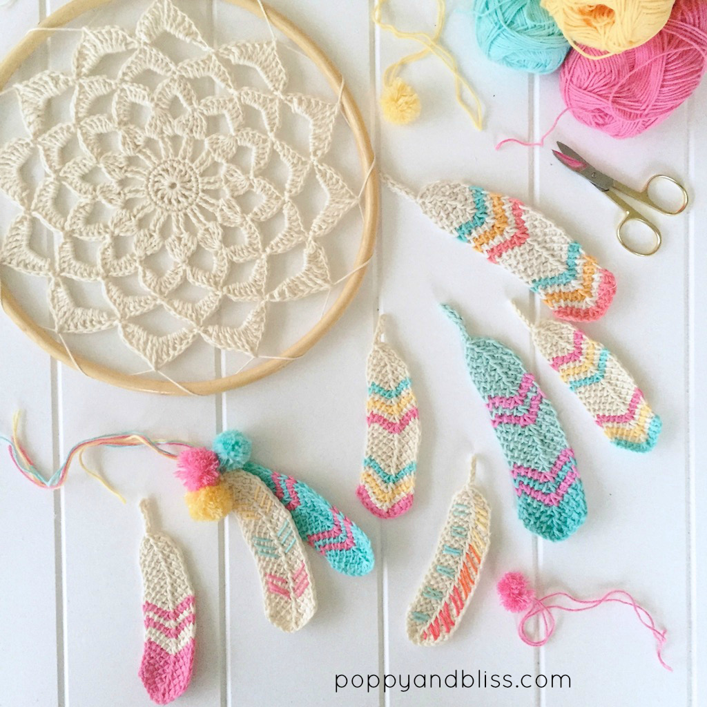 A crochet dreamcatcher and several vibrant crochet feathers are arranged on a white surface with balls of yarn, scissors, and a yarn needle nearby. The image features the text “poppyandbliss.com” in the corner.