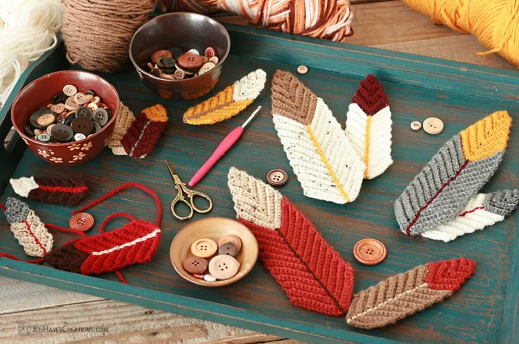 A tray displays colorful crochet feathers, bowls of buttons, balls of yarn, scissors, a crochet hook, and loose buttons—all neatly arranged on a rustic wooden surface.