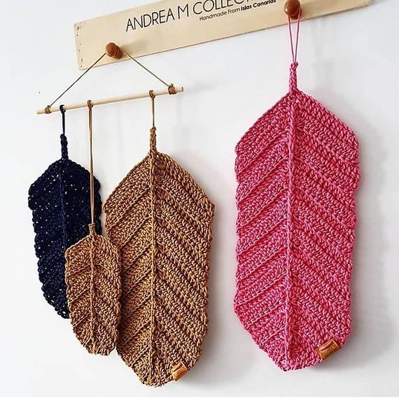 Four large crochet feather leaves in black, brown, and pink hang on a wall; three are attached to a horizontal rod, while one pink leaf hangs separately to the right. A wooden tag above reads ANDREA M COLLECTION.
