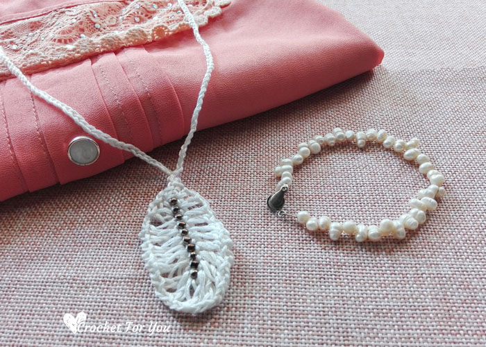 A white crocheted leaf pendant necklace and a beaded pearl bracelet are displayed on pink folded fabric with lace trim and a white button, alongside crochet feather accents. The text Crochet For You is in the bottom left corner.