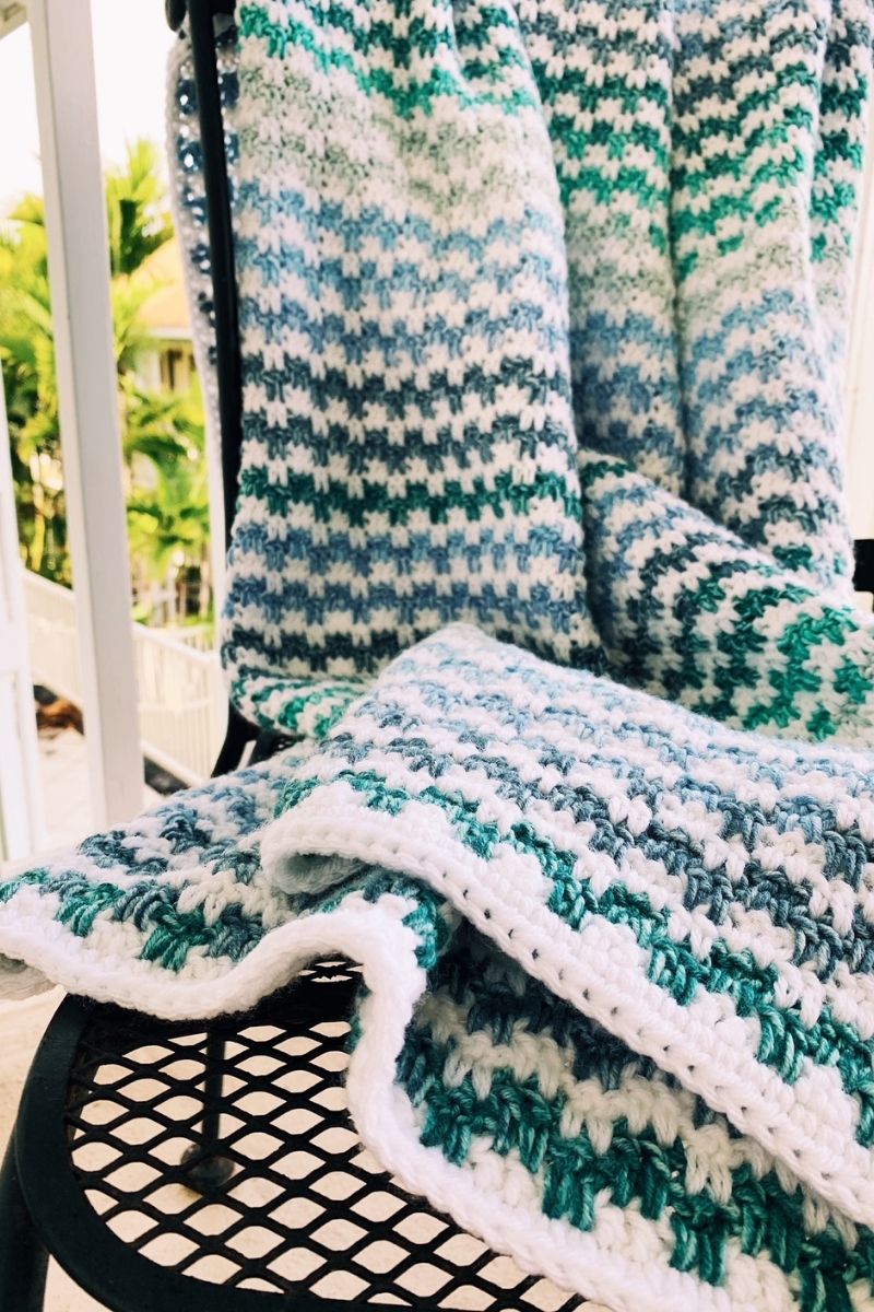 A close-up of a crochet blanket with a white, teal, and light blue pattern draped over a black metal chair with a mesh seat on an outdoor porch. Trees and a white railing are visible in the background.