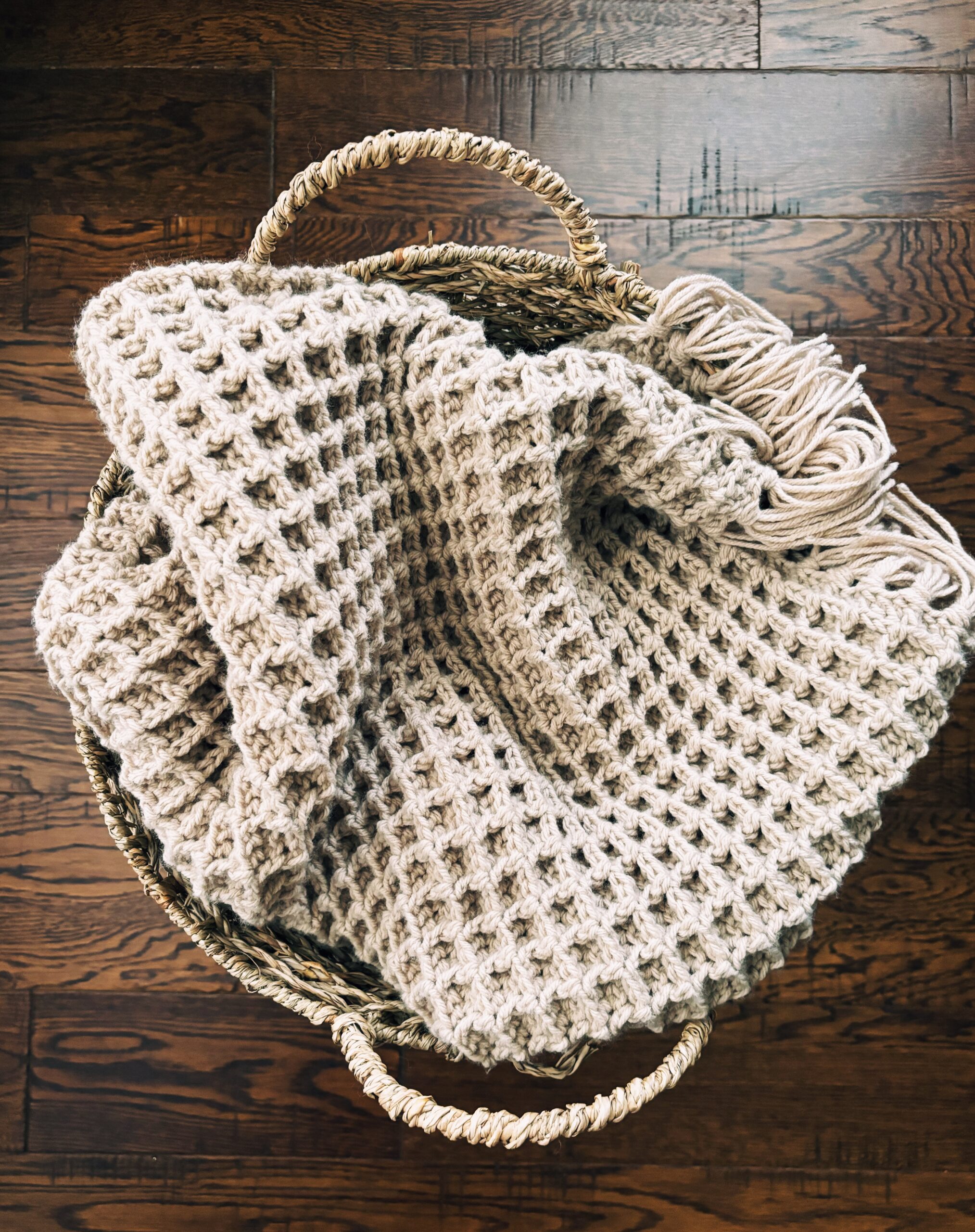 A beige, chunky waffle-knit blanket with fringes is folded in a round, woven basket on a dark wooden floor, reminiscent of the cozy texture found in a classic waffle stitch crochet dishcloth pattern.