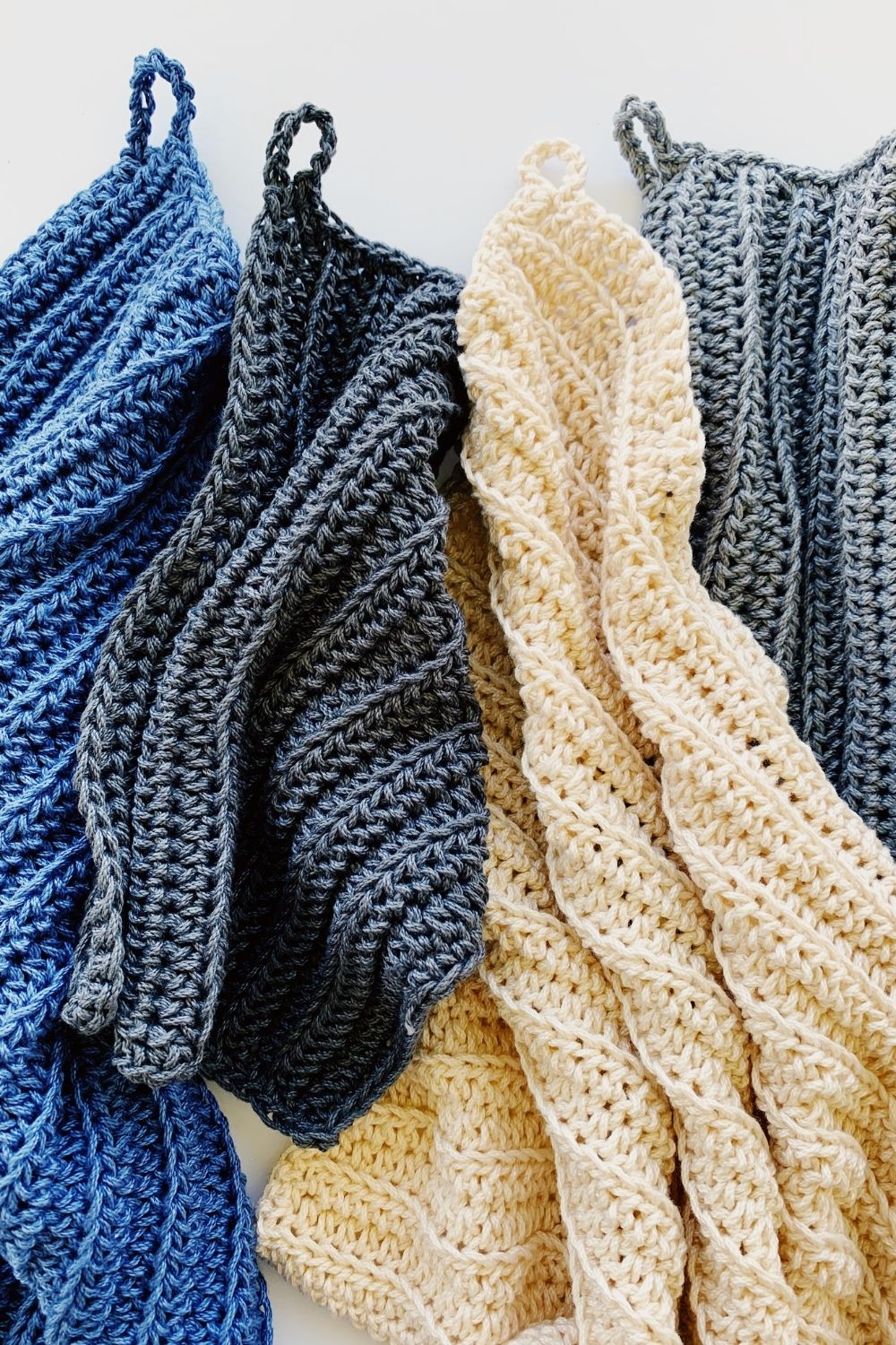 Four textured, ribbed crochet cloths in blue, dark gray, beige, and light gray are spread out on a flat white surface, each beautifully showcasing the waffle stitch crochet dishcloth pattern.