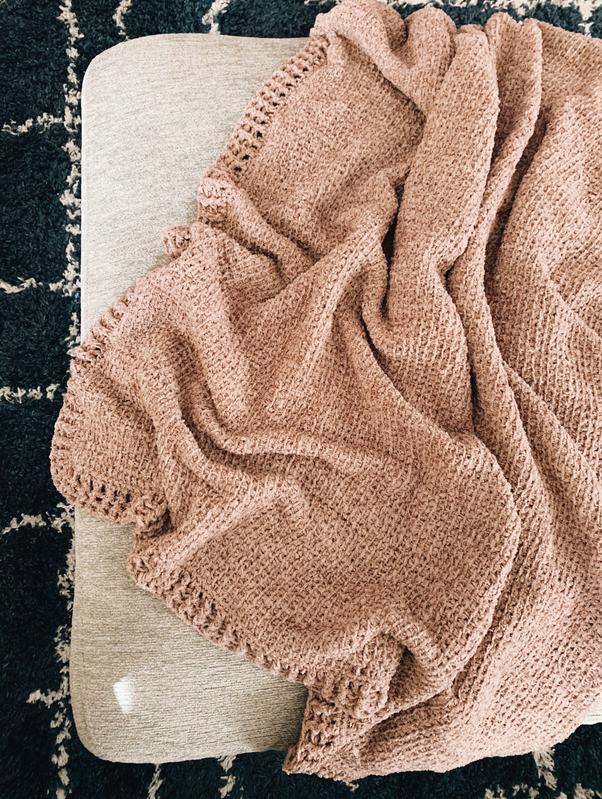 A textured, light brown crochet knit blanket is loosely draped over a beige ottoman, with part of a black and white patterned rug visible underneath.