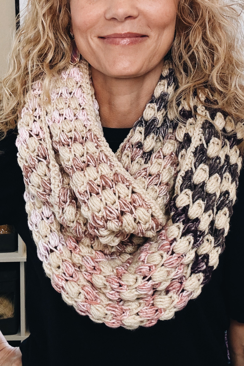 A woman with curly blonde hair wears a chunky crochet infinity scarf in shades of cream, pink, and dark purple over a black shirt. Only her lower face and neck are visible.