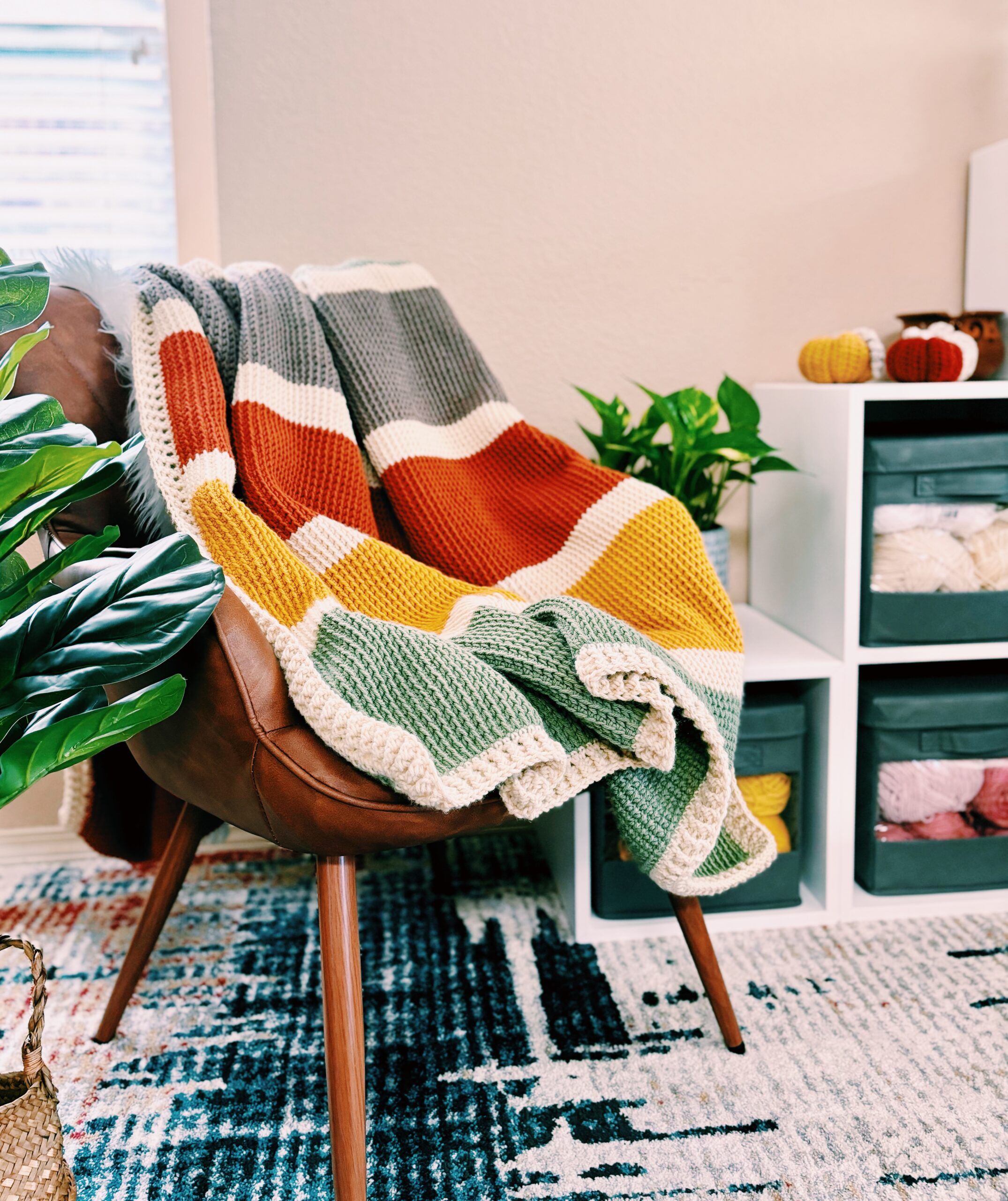 A brown leather chair draped with a colorful crochet blanket sits next to white storage cubes filled with yarn in a cozy, well-lit room with plants and a patterned rug.