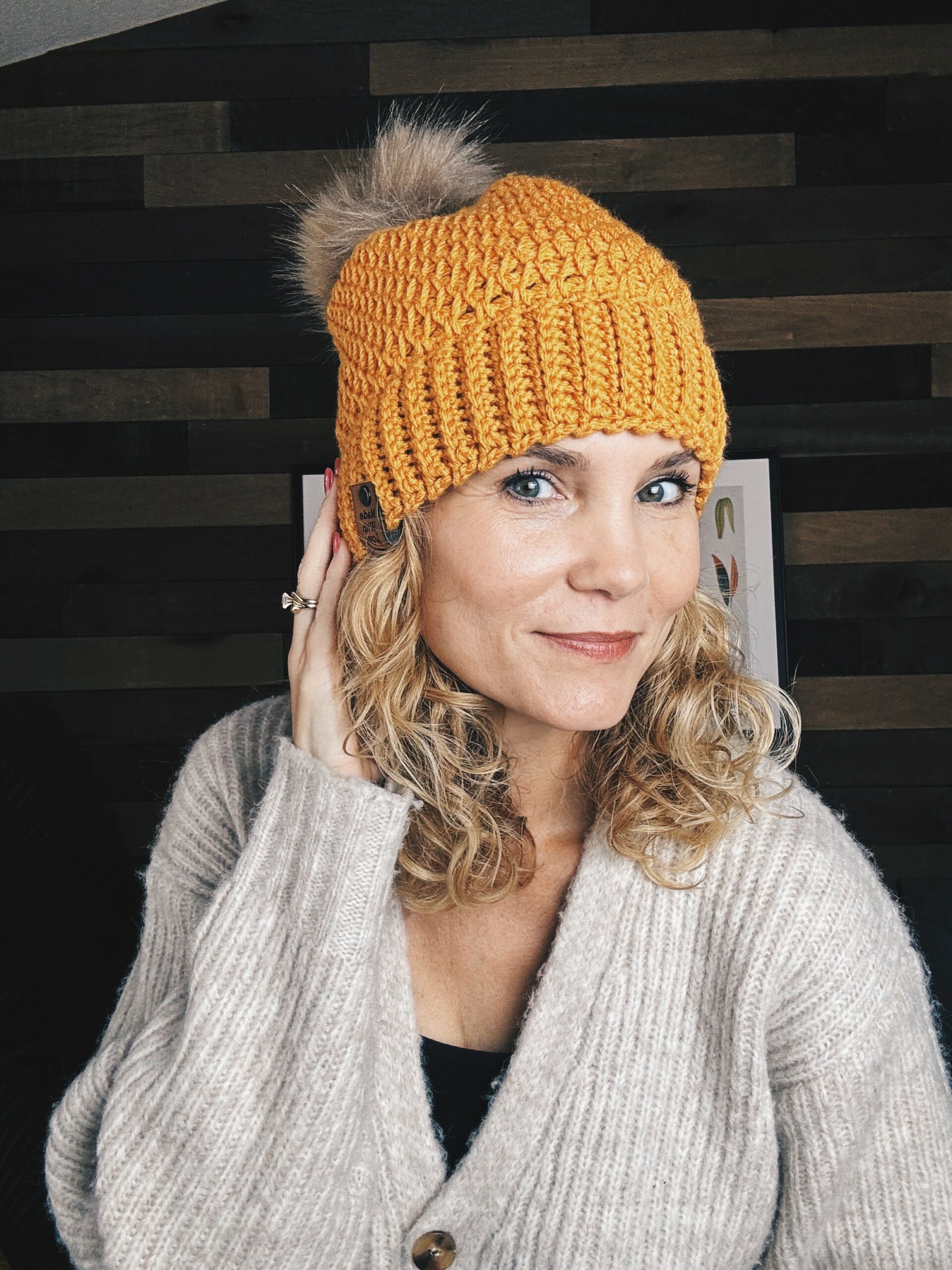 A woman with curly blonde hair wears a yellow knit beanie with a pom-pom and a beige cardigan. She is touching the side of her head and smiling slightly, standing indoors against a dark wooden background.