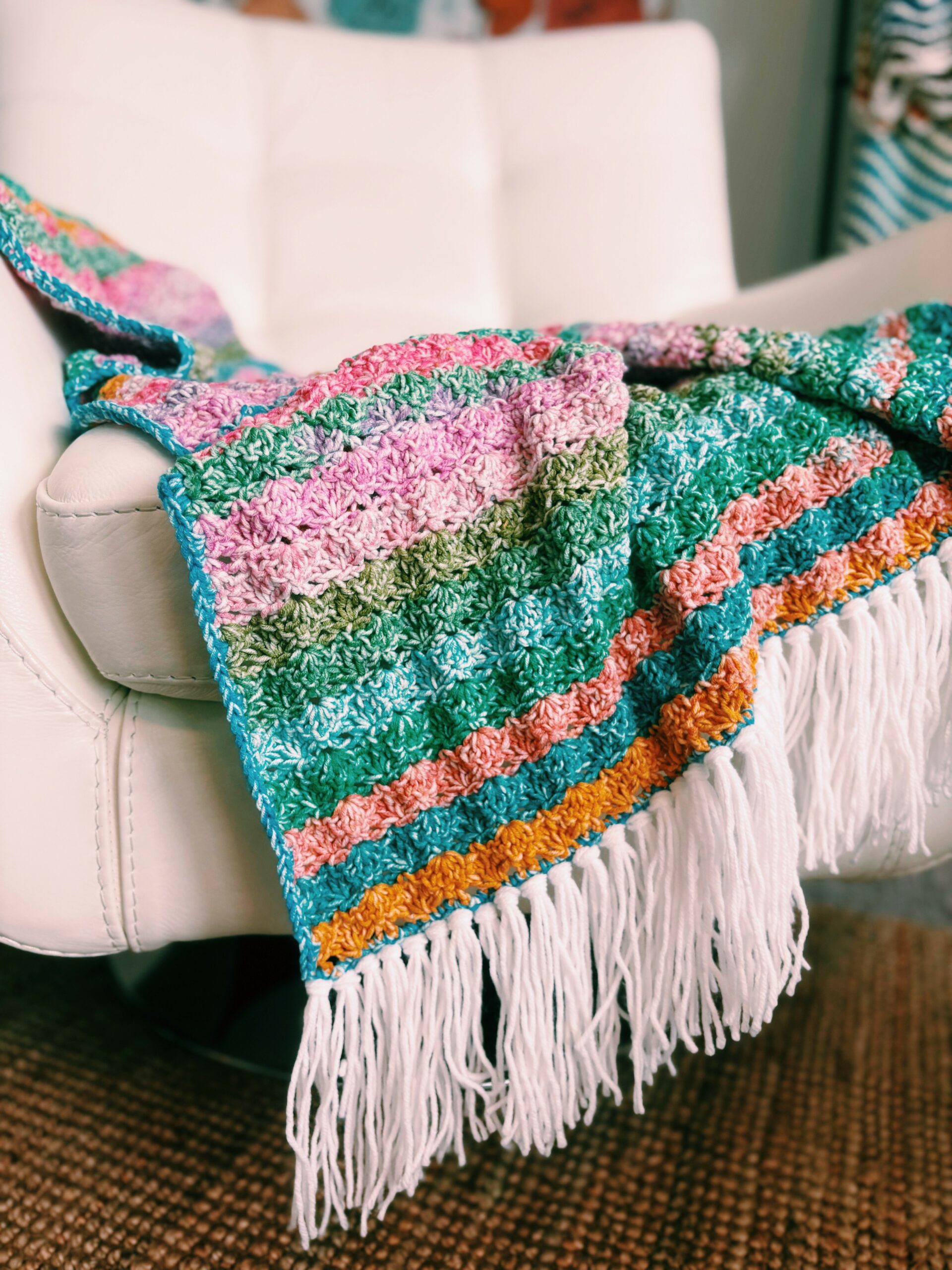 A multicolored crocheted blanket with pink, green, orange, and blue stripes and white fringe is draped over a white cushioned chair. The chair is on a brown woven rug.