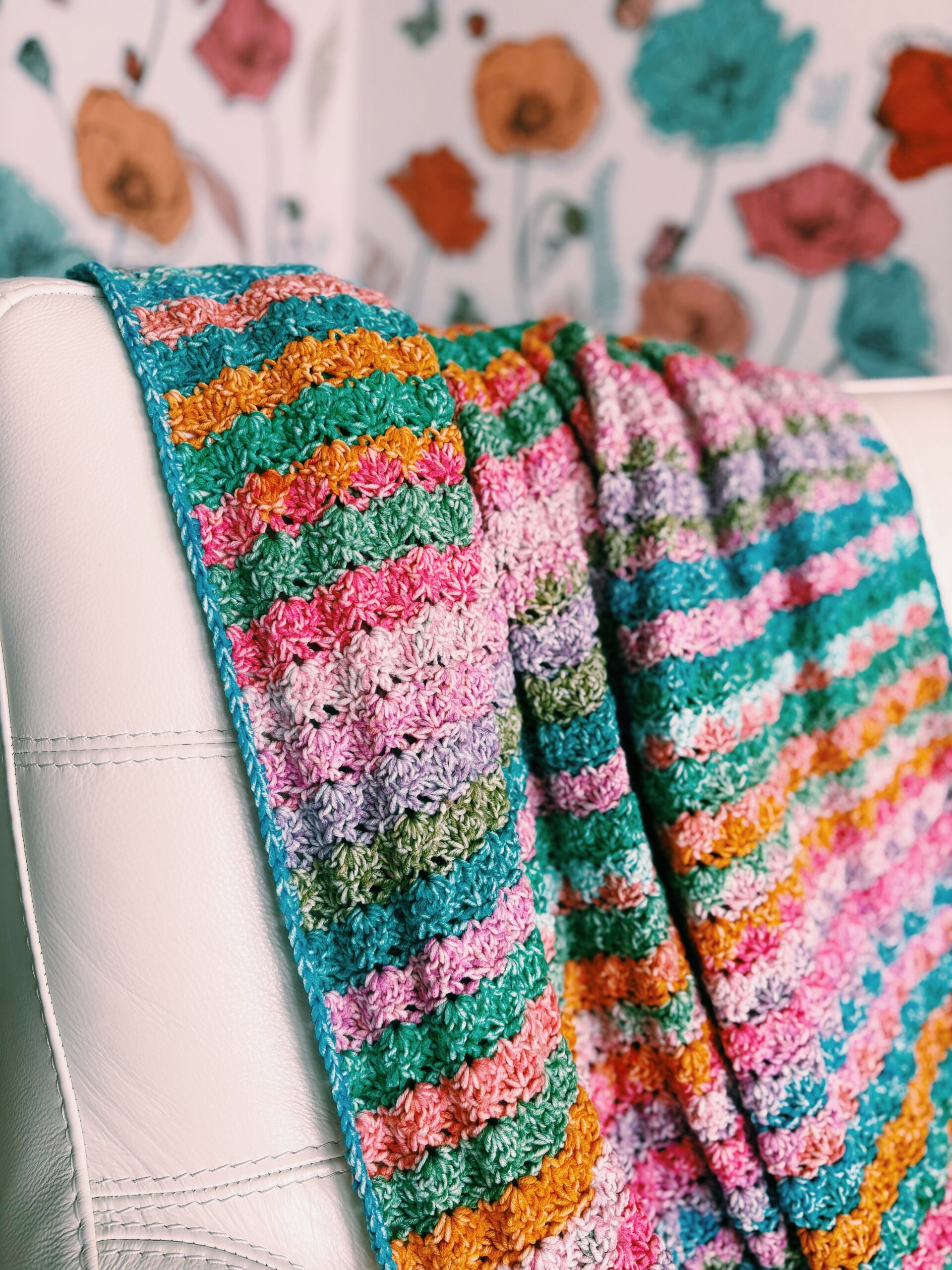A colorful crocheted blanket with striped patterns in pink, green, orange, blue, and purple drapes over the back of a white leather chair. In the background, a wall features a floral poppy pattern.