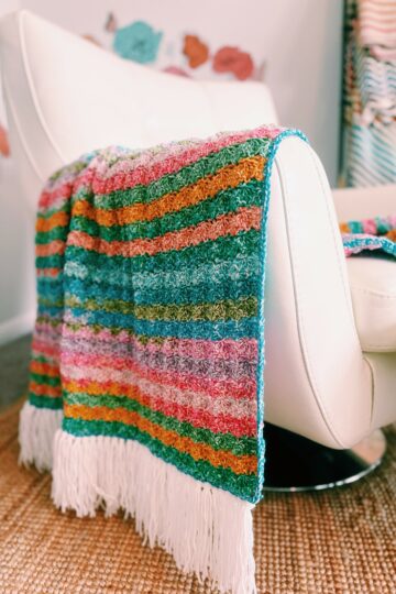A multicolored striped blanket with white fringe is draped over a modern white chair. The background includes another blanket and decorative wall elements. The floor is covered with a woven rug.