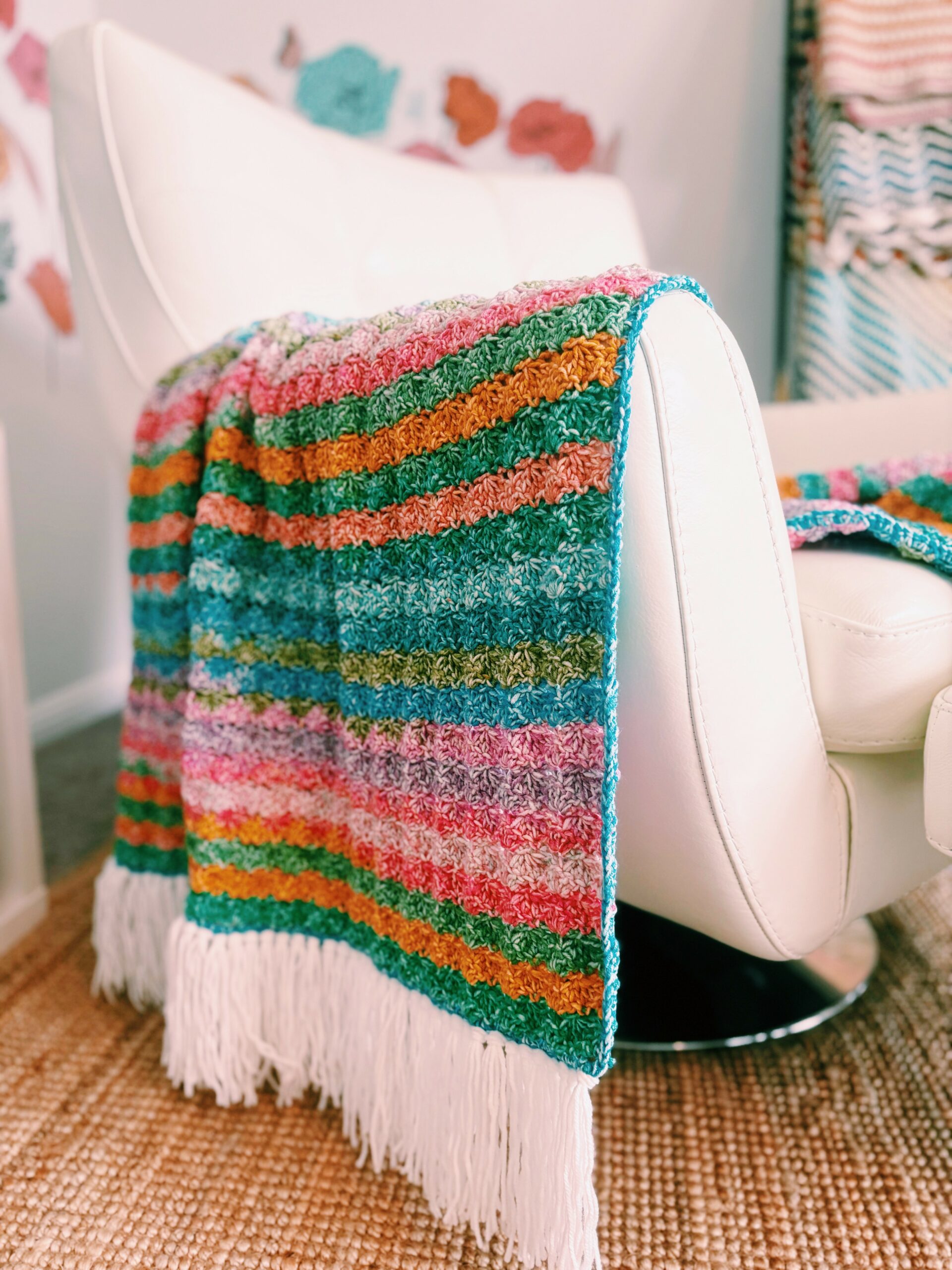 A multicolored striped blanket with white fringe is draped over a modern white chair. The background includes another blanket and decorative wall elements. The floor is covered with a woven rug.