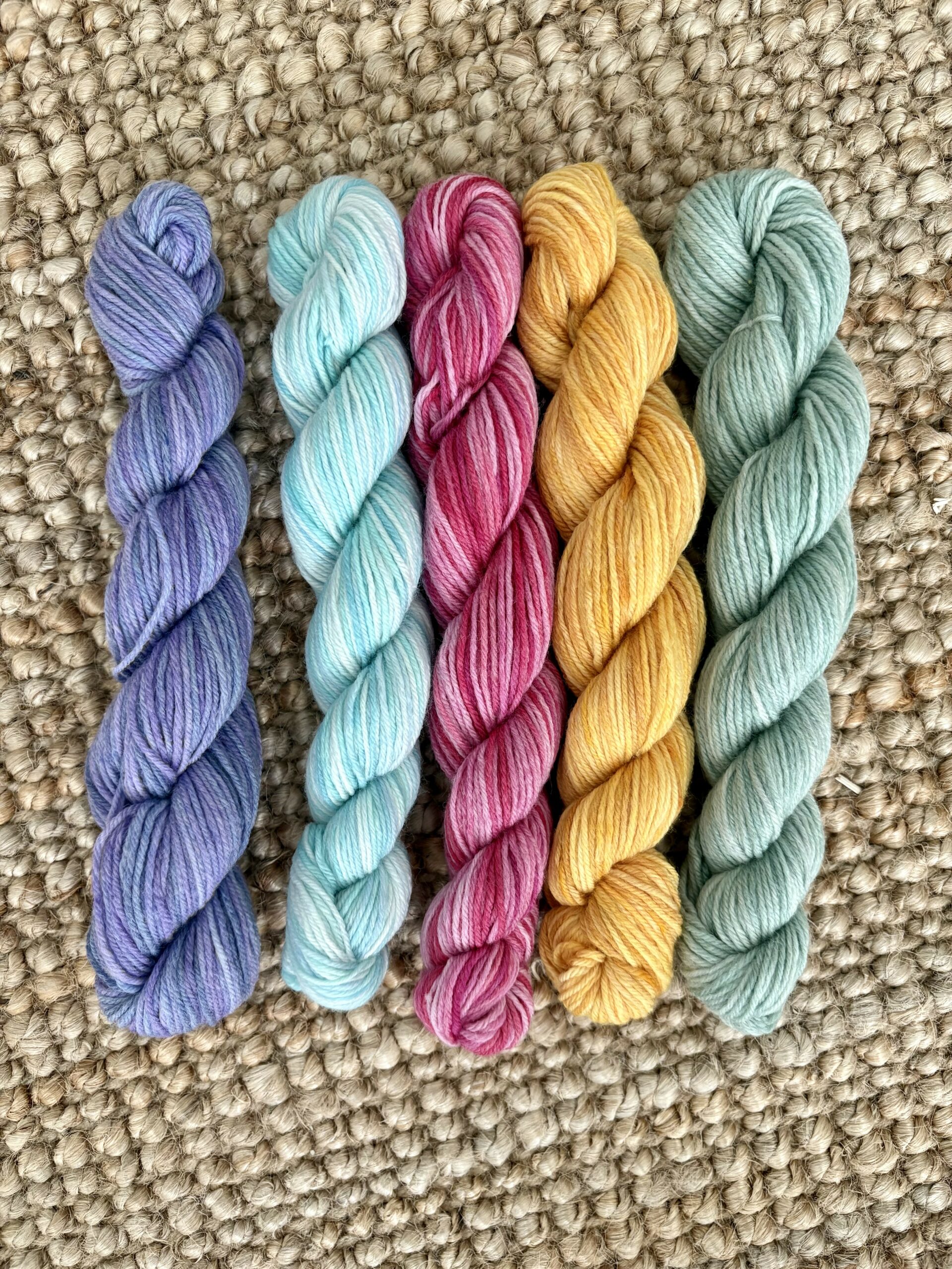 Five skeins of yarn in purple, light blue, pink, yellow, and light green are arranged side by side on a textured beige woven surface.
