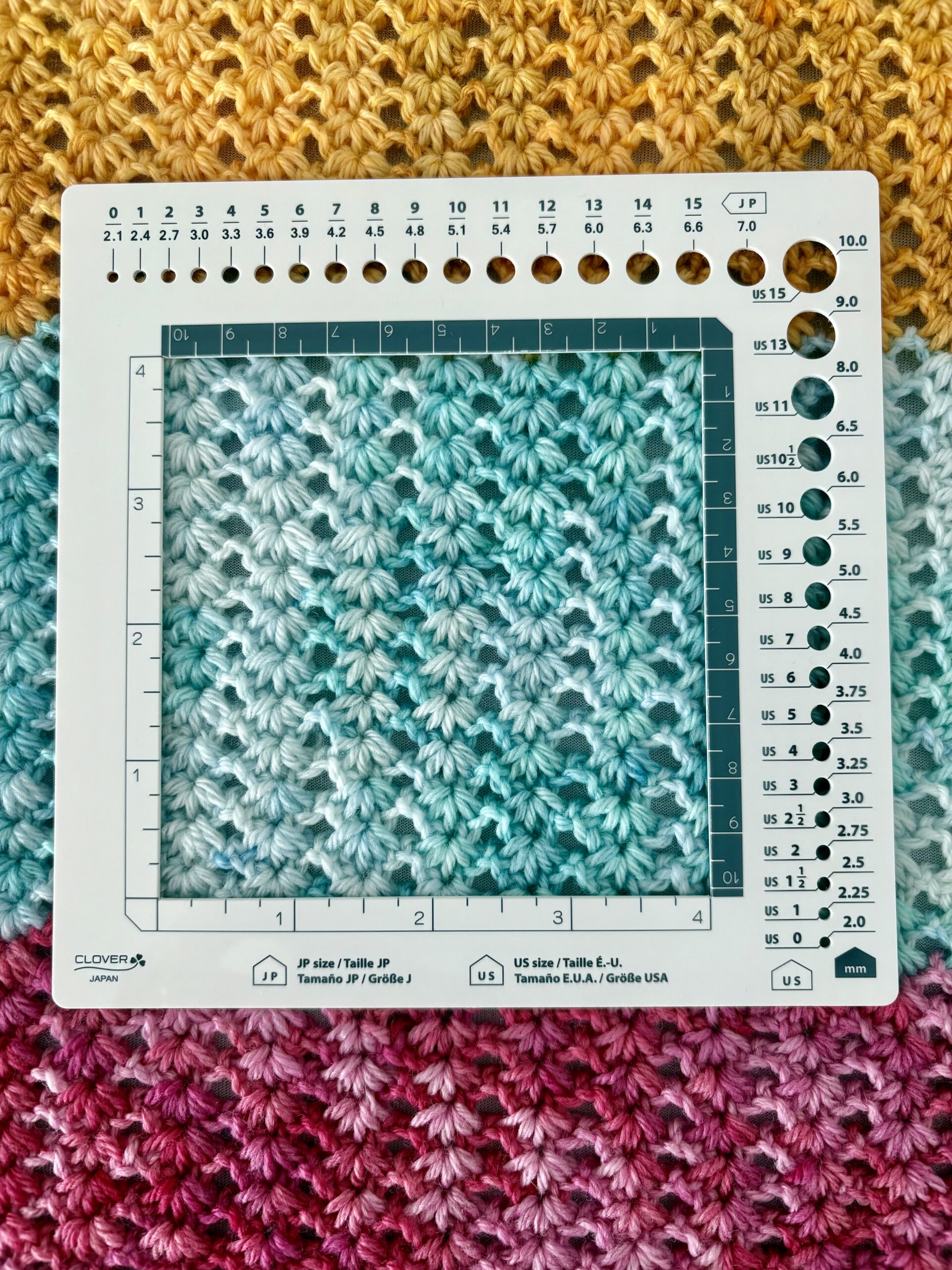 A white knitting and crochet gauge ruler is placed on a multicolored crocheted fabric with yellow, teal, and pink sections in a gradient pattern. The ruler displays measurement units and circular needle size holes.