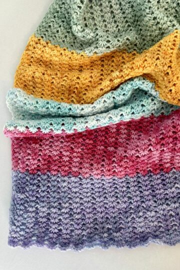 A crocheted fabric with horizontal stripes in green, yellow, light blue, pink, and purple yarn, folded neatly on a light surface.