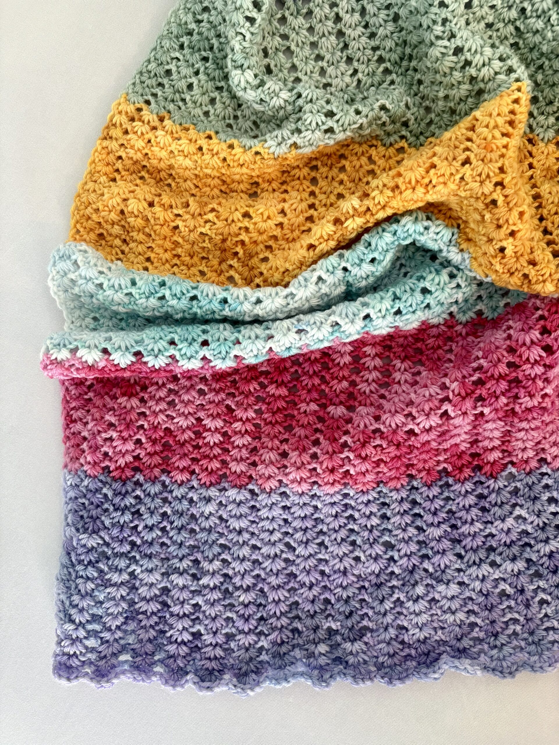 A crocheted fabric with horizontal stripes in green, yellow, light blue, pink, and purple yarn, folded neatly on a light surface.