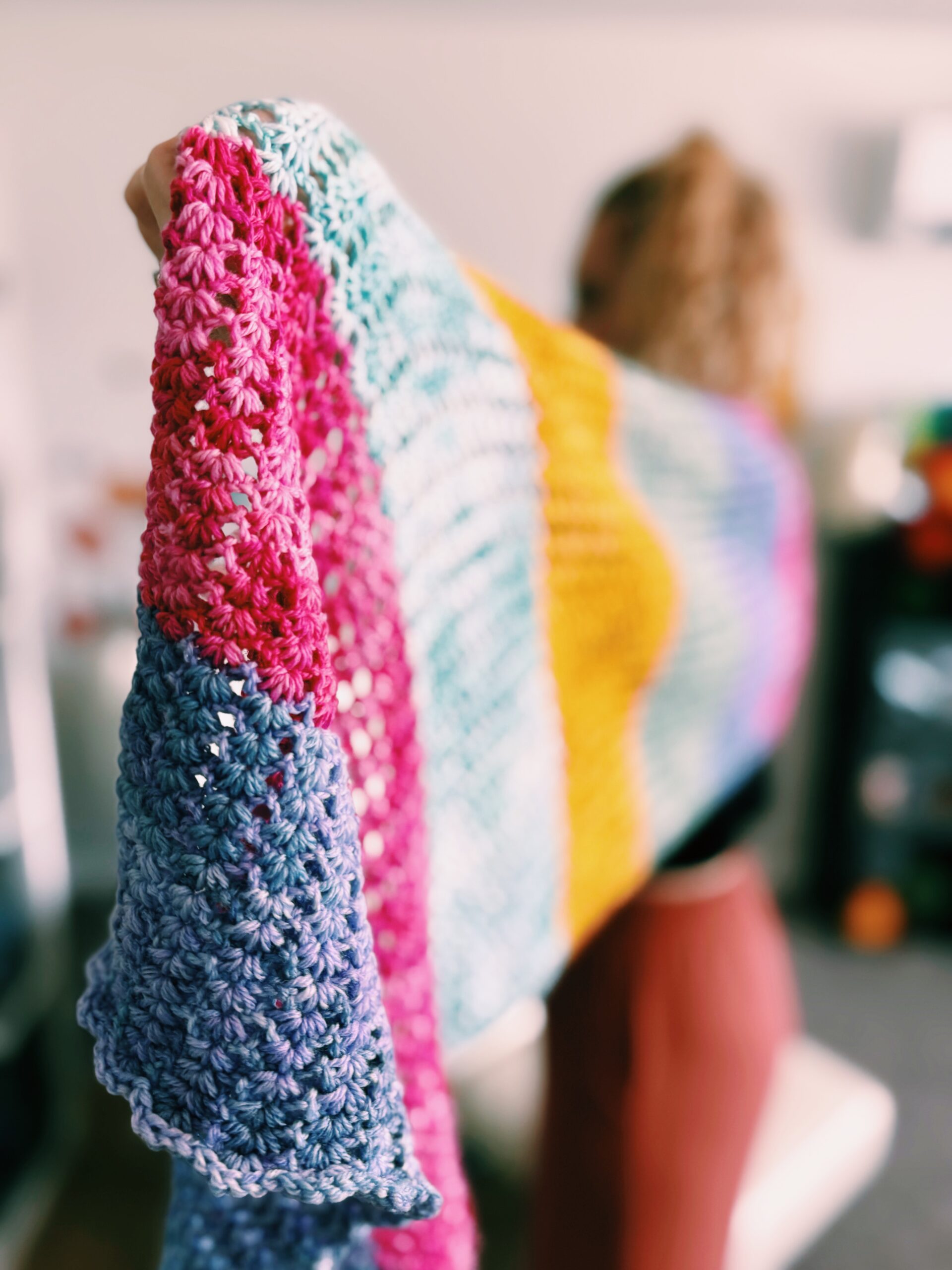 A person holds up a large crocheted blanket with horizontal stripes in blue, purple, pink, green, yellow, and orange tones. The focus is on the blanket; the person is blurred in the background.