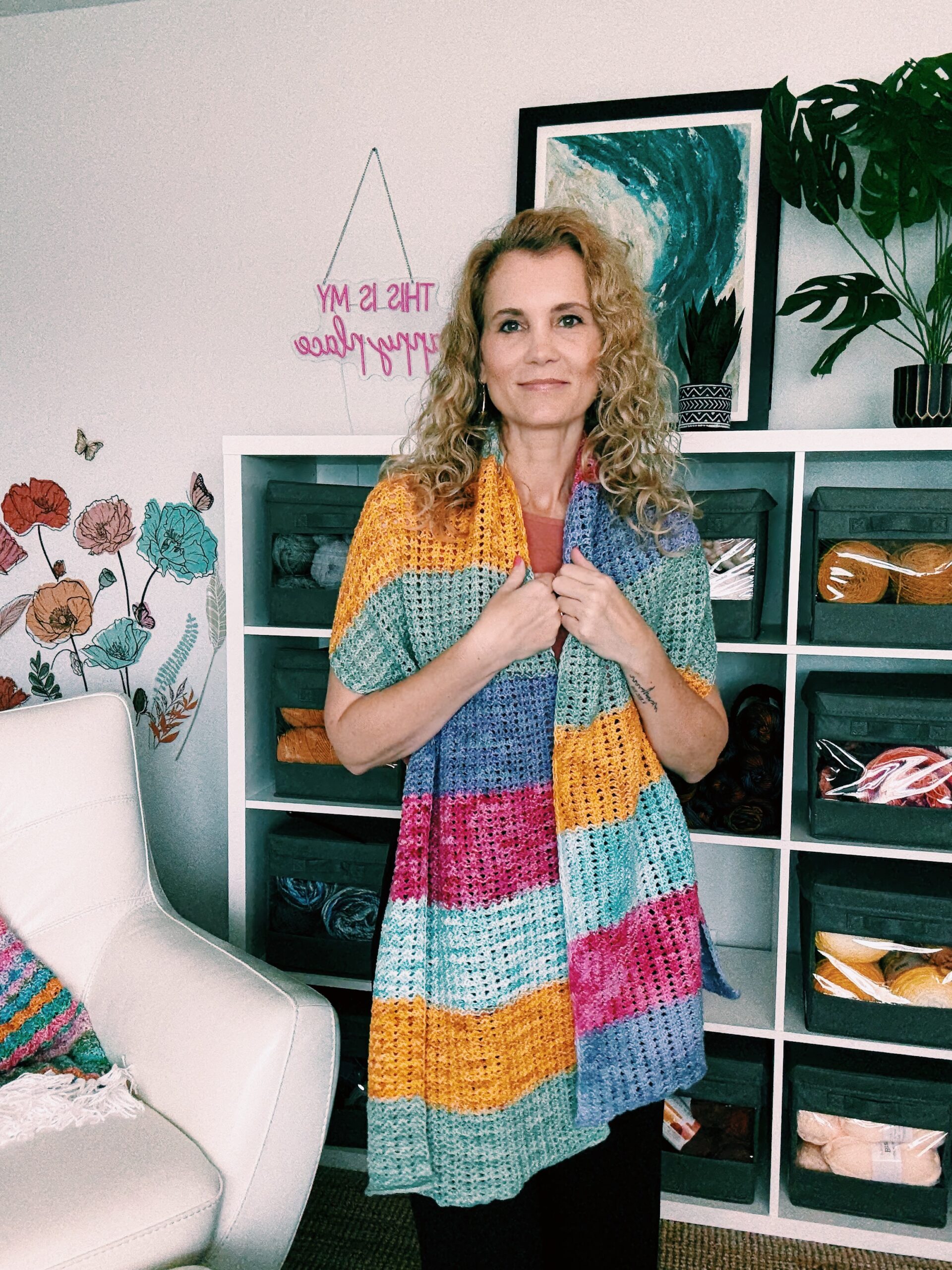 A woman with curly blonde hair stands in front of a yarn shelf, wearing a colorful crocheted shawl. The room is decorated with yarn, a floral wall decal, a white chair, and framed artwork.