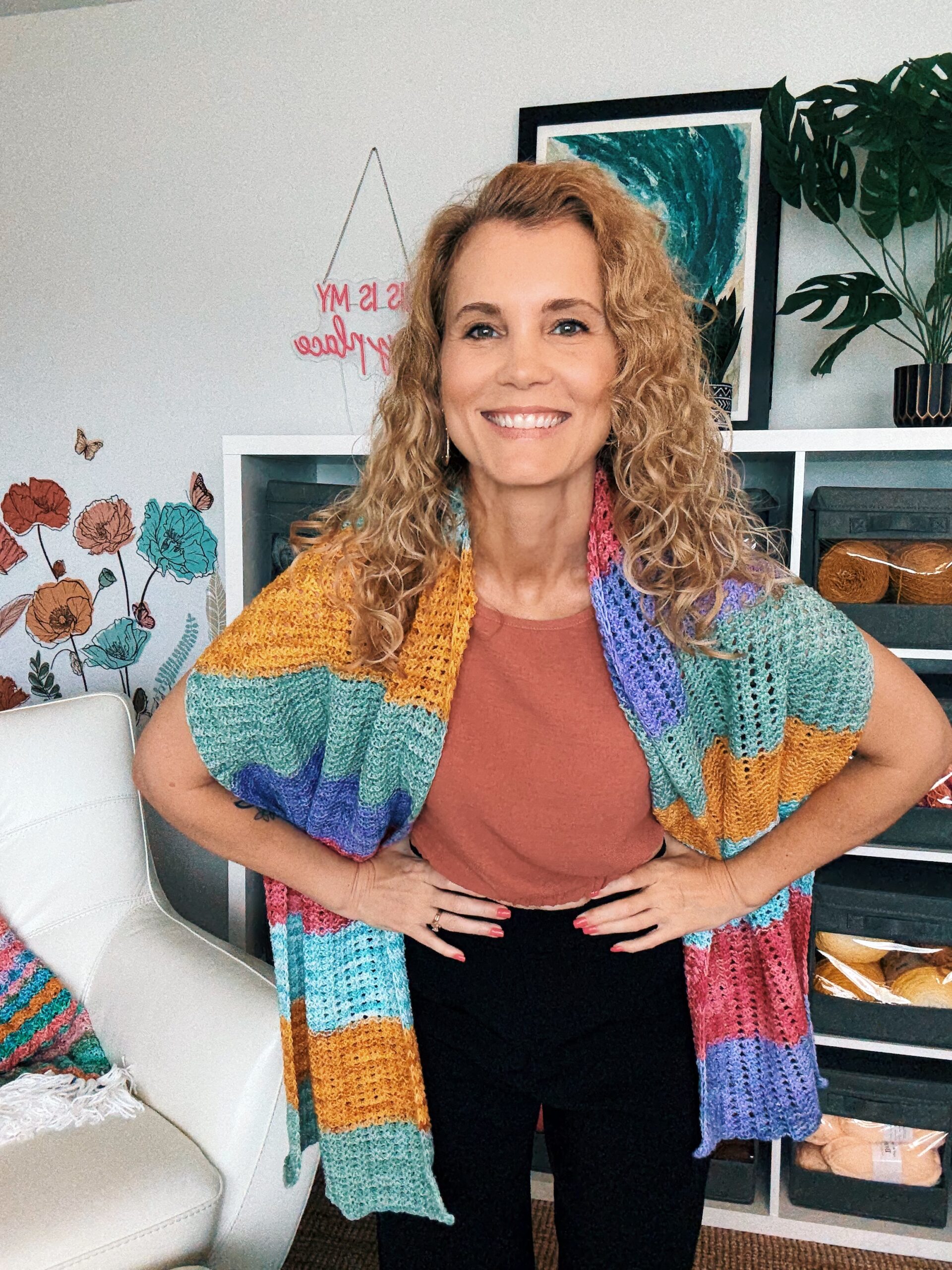 A woman with curly blonde hair smiles at the camera, wearing a rust-colored shirt, black pants, and a colorful striped crochet shawl, standing in a room decorated with plants, yarn, and floral wall art.