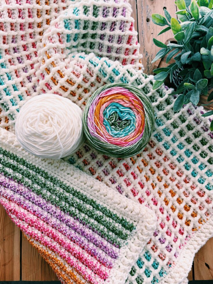 A crochet waffle stitch baby blanket with a colorful pattern and two balls of yarn—one white, one multicolored—rests on a wooden surface next to a green potted plant.