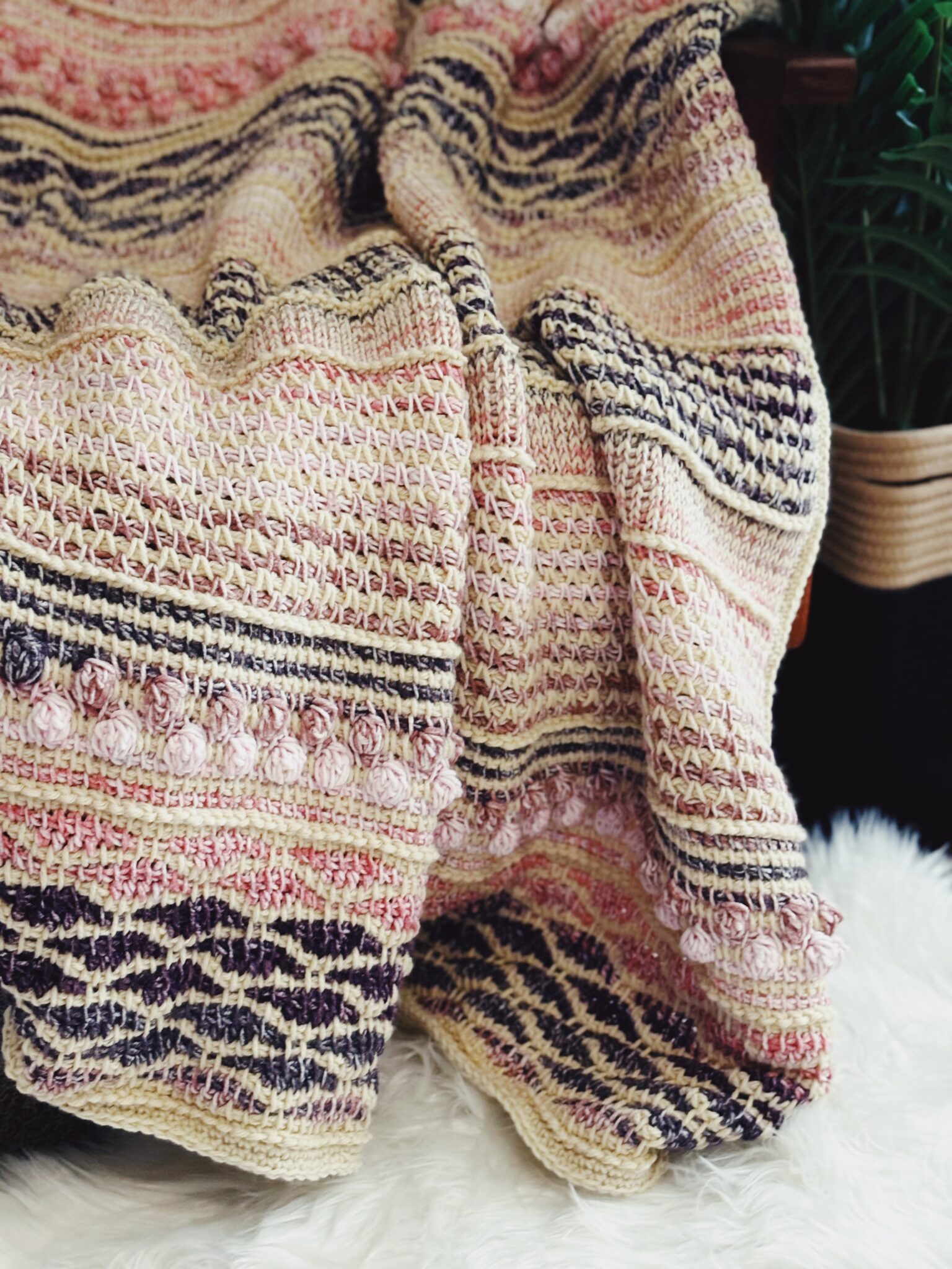 A crocheted blanket with intricate crochet patterns in shades of cream, pink, and dark purple—made with Mandala Ombre yarn—is draped over a surface, complemented by a green potted plant and a white textured rug in the background.
