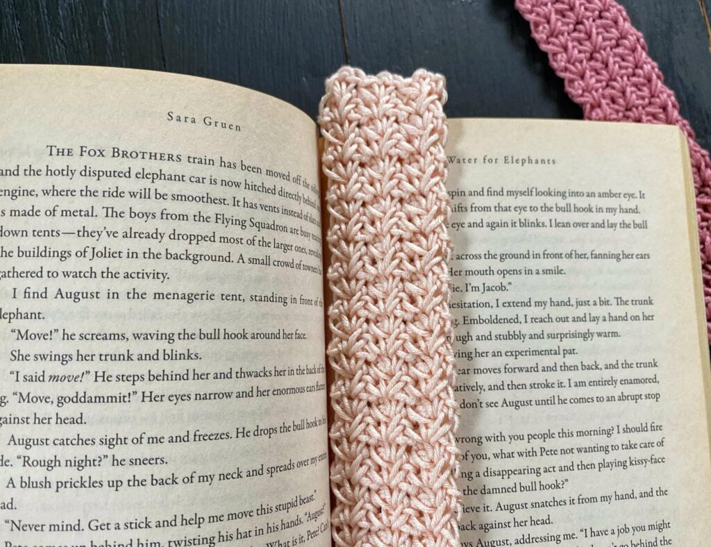 An open book with a crocheted pink and white bookmark placed vertically across the center, offering September cozy vibes and crochet project ideas, with a matching strap visible in the background.
