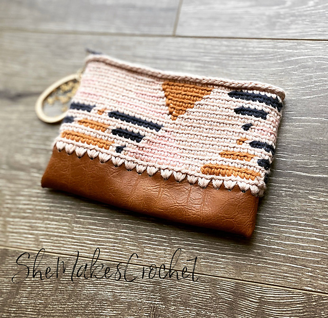 A small pouch with a geometric crochet pattern in pink, brown, black, and white yarn, featuring a brown faux leather bottom—perfect for September cozy vibes or as one of your back to school crochet project ideas, displayed on a wooden surface.