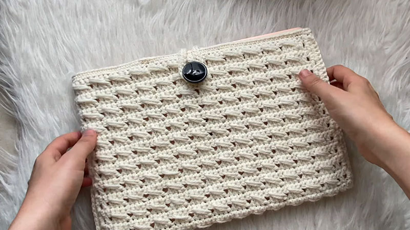 Two hands hold a beige, crocheted pouch with a button closure—an ideal back-to-school accessory or inspiration for your next September crochet project idea—resting on a soft, light-colored surface.