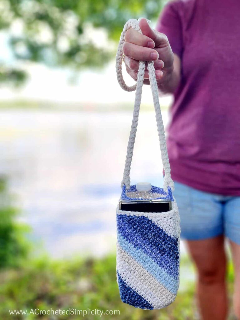 A person in a purple shirt and denim shorts holds a crocheted water bottle holder with a blue and white diagonal stripe pattern—perfect for September Cozy vibes or as inspiration for Back-to-School Crochet project ideas.