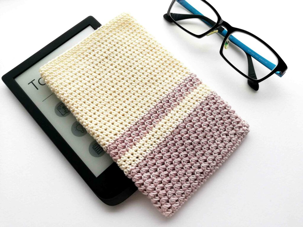 A black e-reader, partially covered by a crocheted sleeve with cream and lavender stripes—perfect for September Cozy vibes—rests beside black eyeglasses with blue temples.