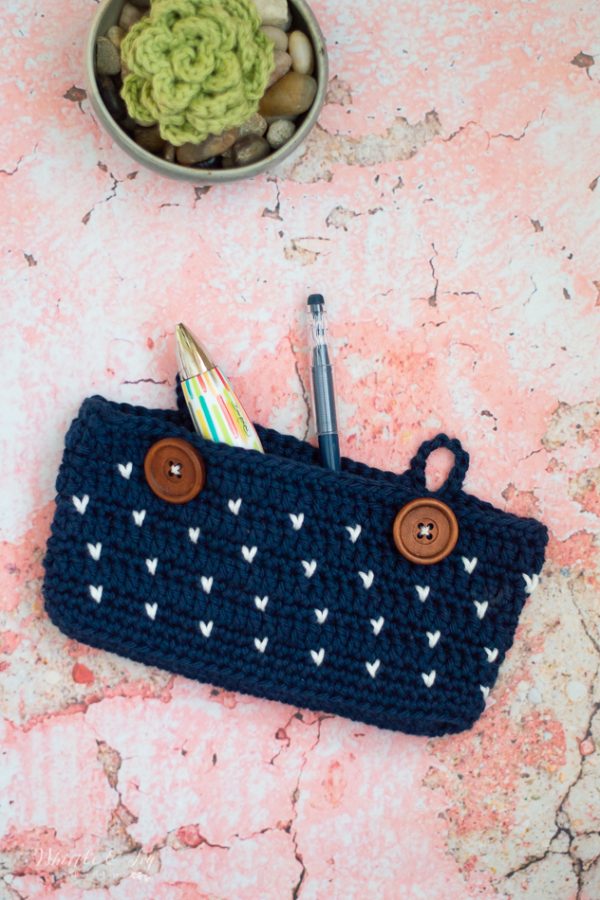 A blue crocheted pouch with wooden buttons holds a pen and a colorful marker—perfect for Back-to-School organization. Above, a small bowl with a crocheted flower and rocks sits on a pink marbled surface, inspiring new Crochet Project Ideas for September.