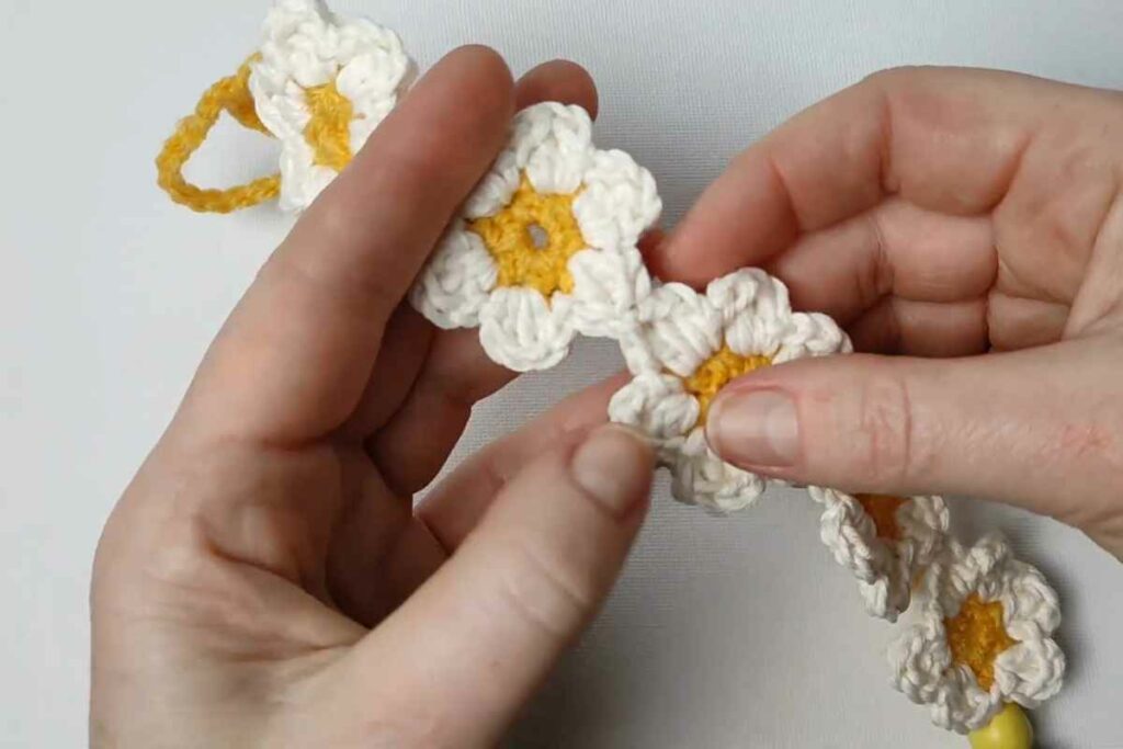 Two hands hold a crocheted flower chain with white petals and yellow centers—a charming inspiration for September crochet or back-to-school crochet project ideas—against a plain background.