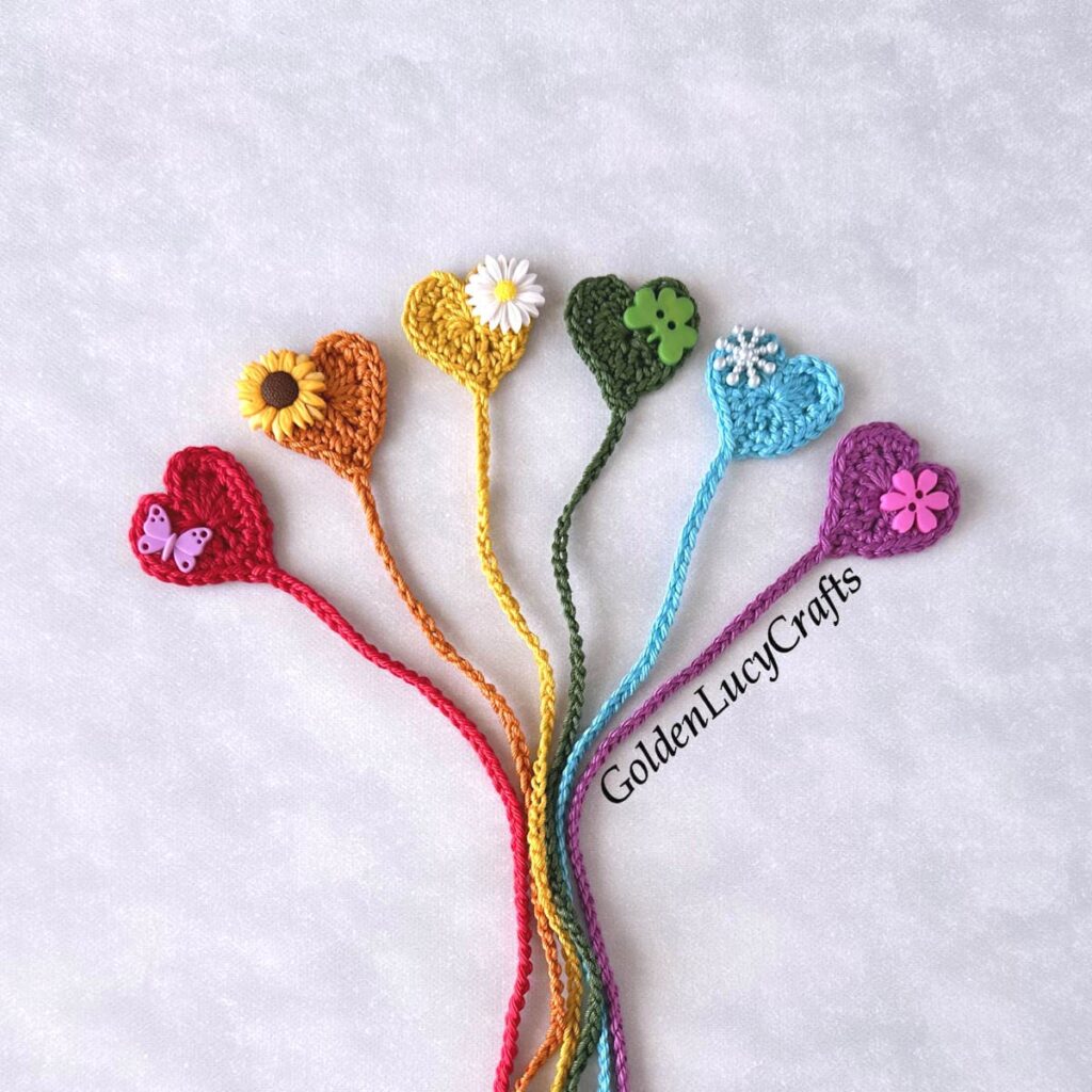 Six crocheted heart-shaped bookmarks in different colors, adorned with flower and butterfly embellishments, are arranged in a fan shape on a light background—perfect for Back-to-School crochet or September cozy crochet project ideas.