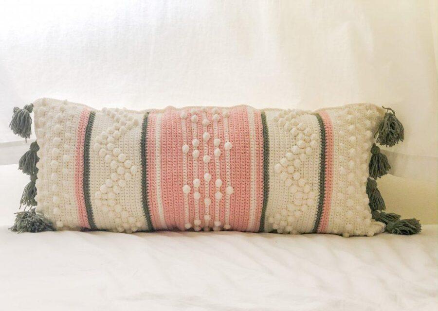 A rectangular decorative pillow with textured patterns in white, pink, and green, featuring tassels on each corner, rests on a white surface—a cozy crochet accent perfect for fresh crochet project ideas.