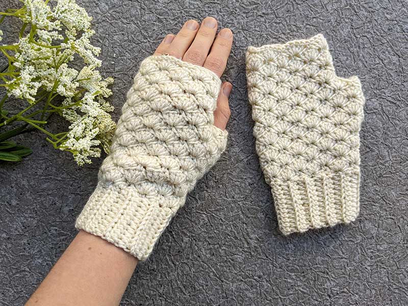 A hand wearing a cream-colored, crocheted fingerless glove with another matching glove beside it, on a gray textured surface next to white flowers—perfect for September cozy vibes and back-to-school crochet inspiration.