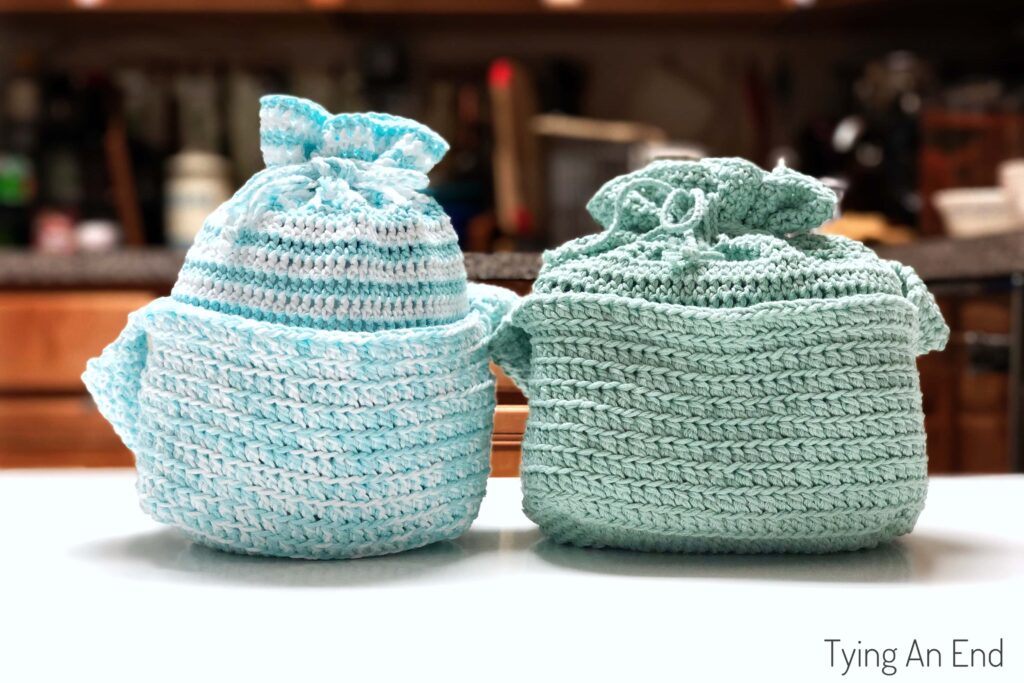 Two light blue crocheted drawstring bags sit side by side on a white surface, adding a touch of cozy crochet charm to your space, with a blurred kitchen background.