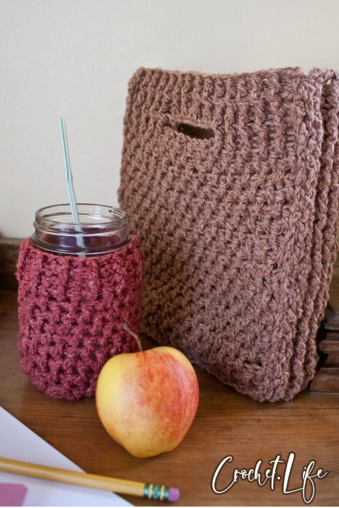 A pink mason jar cozy with a crochet hook inside, a red and yellow apple, and a brown crocheted bag sit on a wooden surface—perfect September inspiration for back-to-school crochet project ideas.