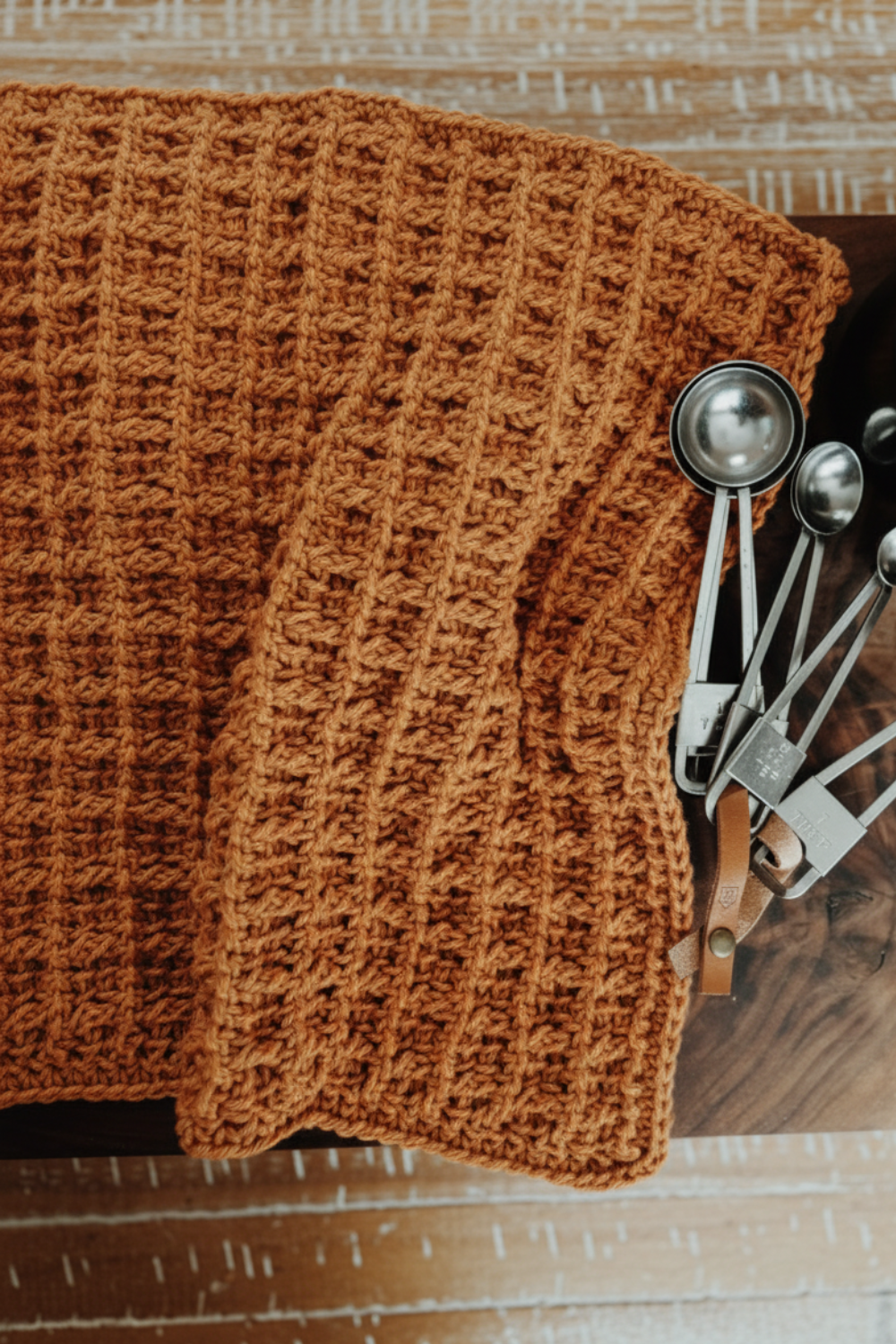 A brown knitted blanket with a waffle pattern is draped over a surface next to a set of metal measuring spoons.