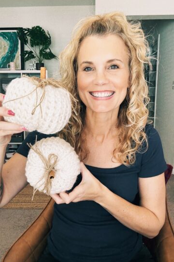 A woman with curly blonde hair smiles while holding two white knitted pumpkins with twine bows in a cozy home setting, perfect for showcasing a new free pumpkin pattern.