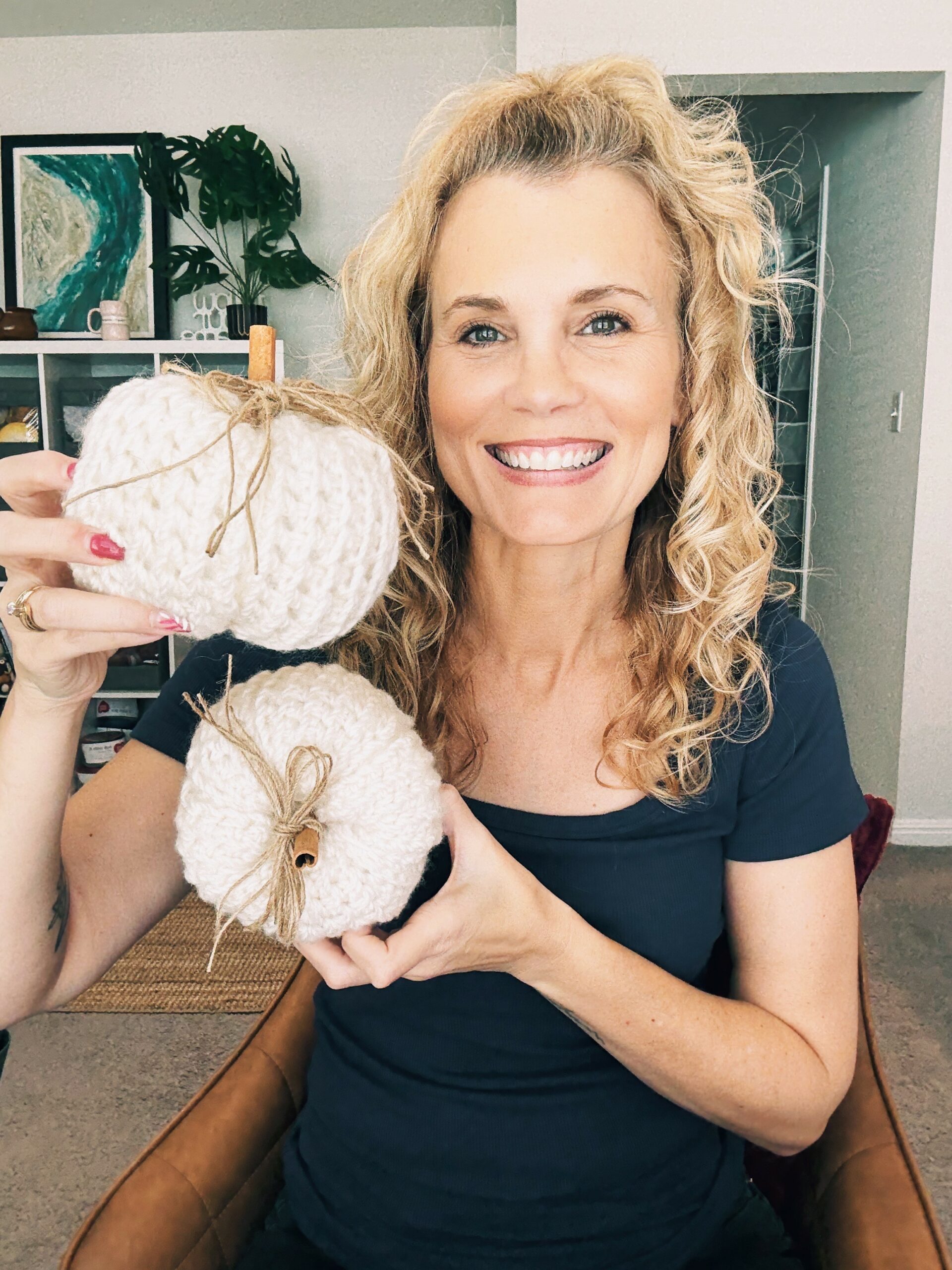 A woman with curly blonde hair smiles while holding two white knitted pumpkins with twine bows in a cozy home setting, perfect for showcasing a new free pumpkin pattern.