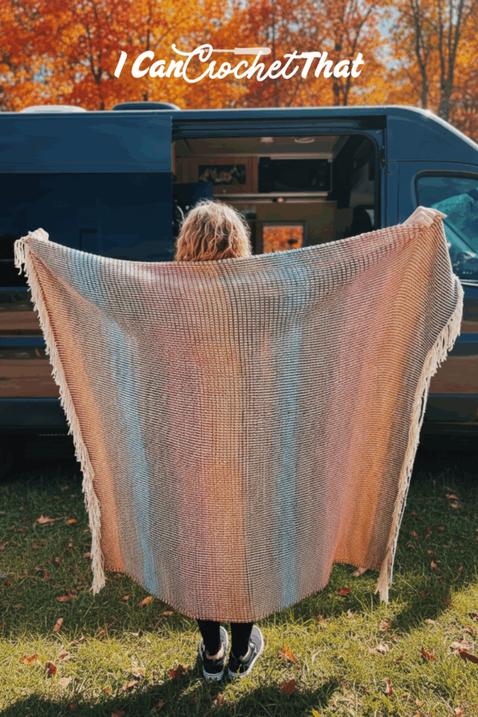 Person holding a large, multicolored crocheted blanket in front of a blue van, with autumn trees in the background. The words I Can Crochet That appear at the top.