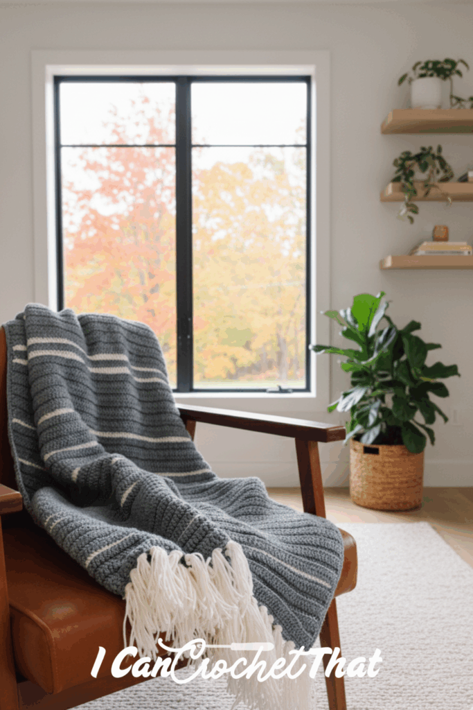 A striped gray and white crochet blanket draped over a brown armchair in a bright room with a window, potted plant, and floating shelves; text reads I Can Crochet That.