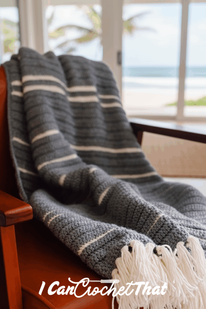 A gray and white striped crochet blanket with tassels draped over a brown chair, with a beach and palm trees visible through the window in the background.