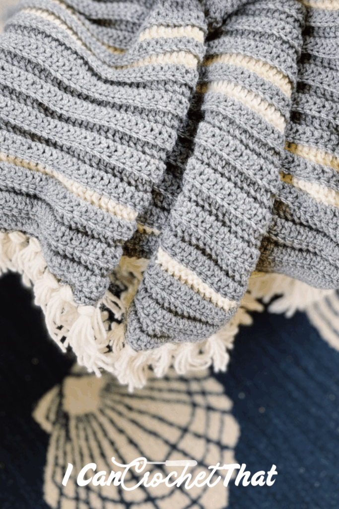 A folded gray and cream striped crochet blanket with fringe edges is displayed on a navy surface with shell designs.