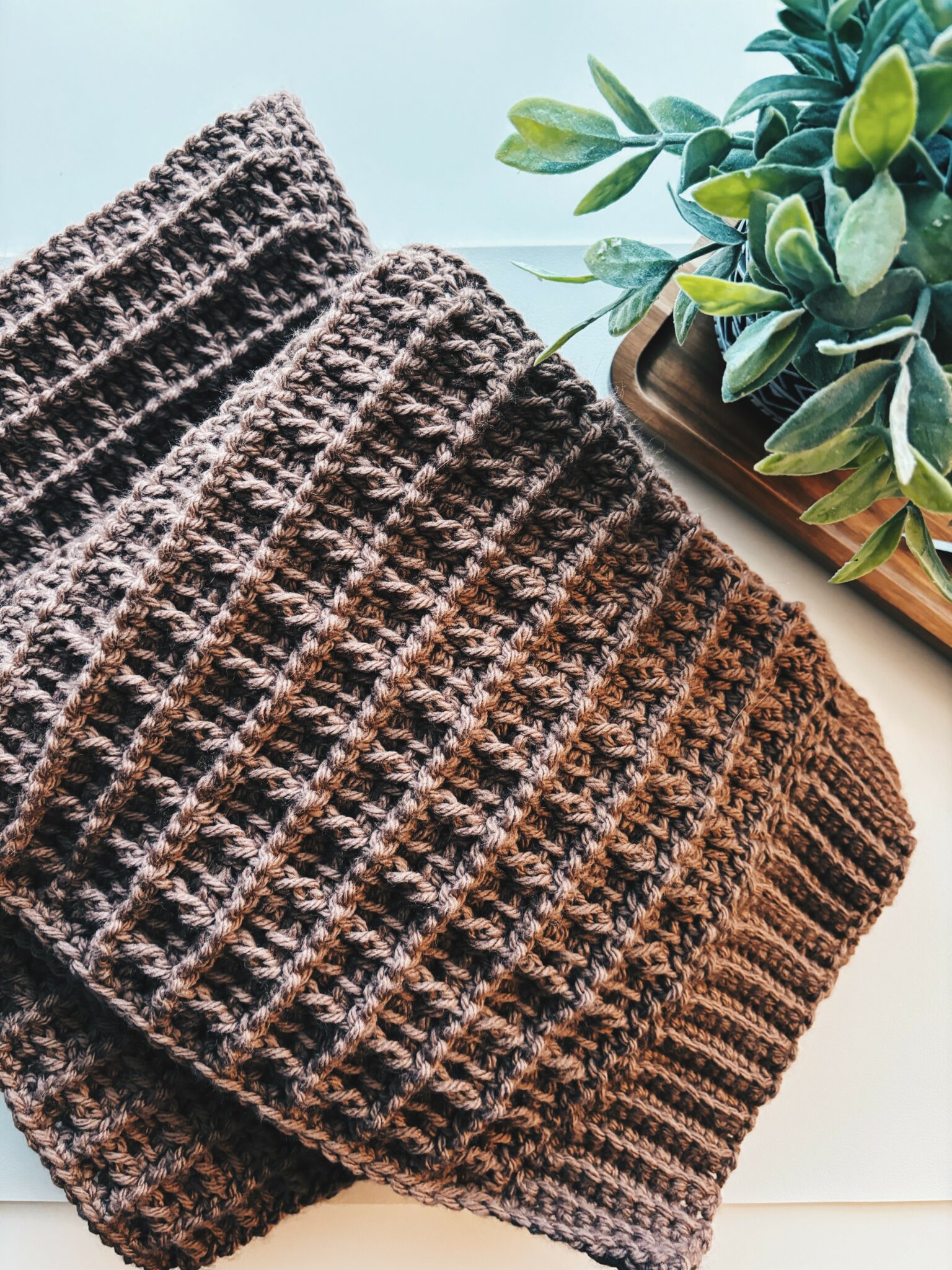 A folded brown knitted blanket, crafted with a Tunisian Waffle Stitch, sits on a white surface next to a potted green leafy plant in a wooden tray.