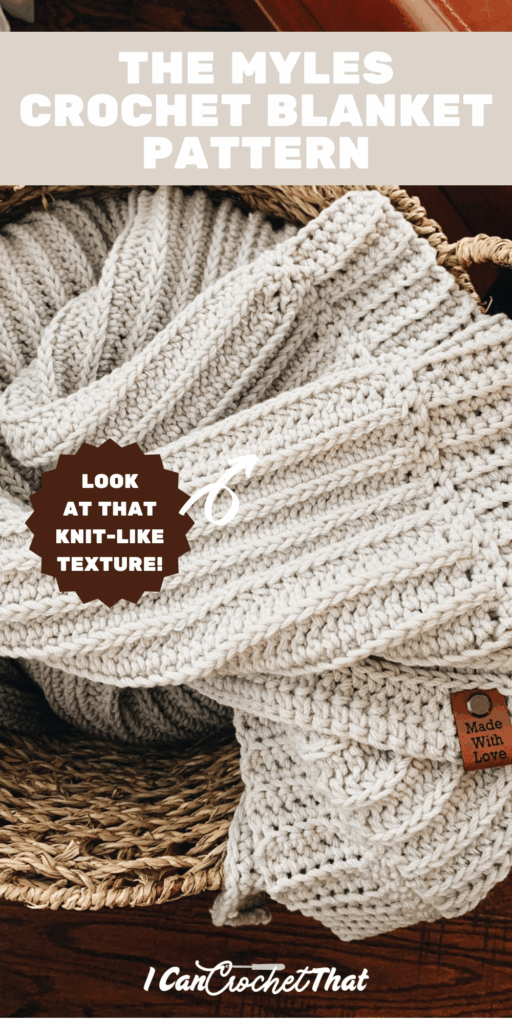 A cream-colored crochet blanket with a knit-like texture is folded in a woven basket. The blanket has a small leather tag labeled Made With Love.