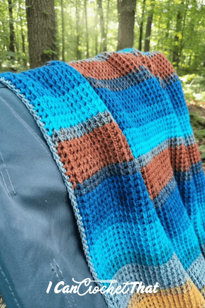 A colorful blue, brown, and gray crocheted blanket draped over a chair outdoors in a forest setting. The text I Can Crochet That appears at the bottom.