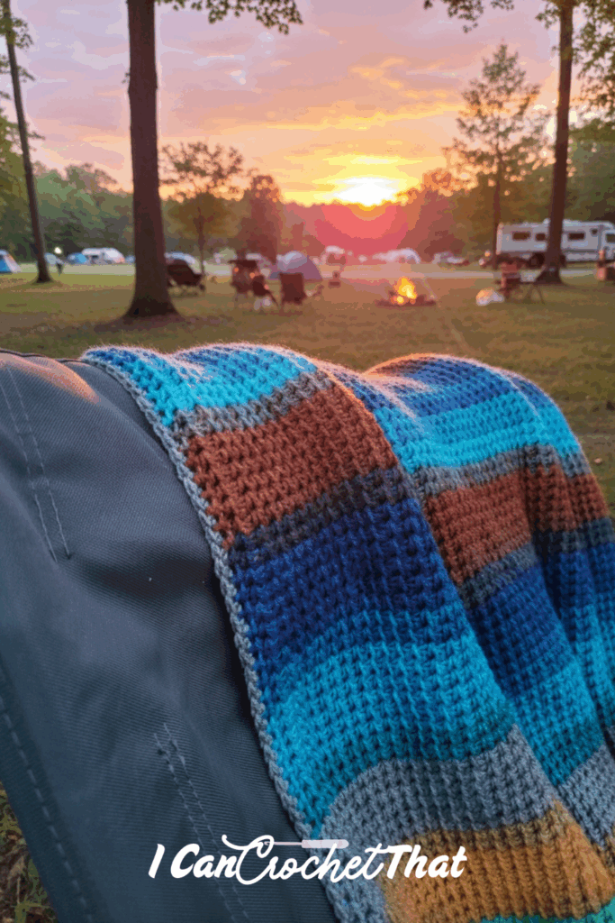 A crocheted blanket draped over a chair at a campsite with tents, RVs, a campfire, and a sunset in the background. Text at the bottom reads I Can Crochet That.