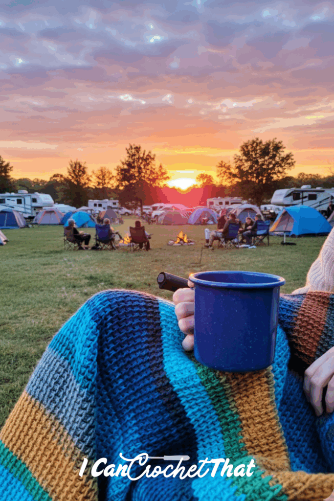 Person holding a blue mug and striped crochet blanket at a campsite with tents, RVs, a fire pit, and a sunset in the background.