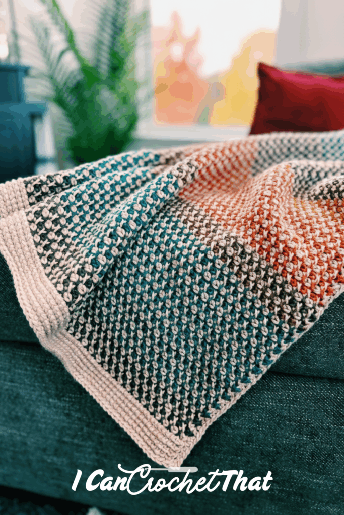 A textured crochet blanket in blue, white, and orange tones is draped over a dark sofa. The logo I Can Crochet That appears at the bottom.