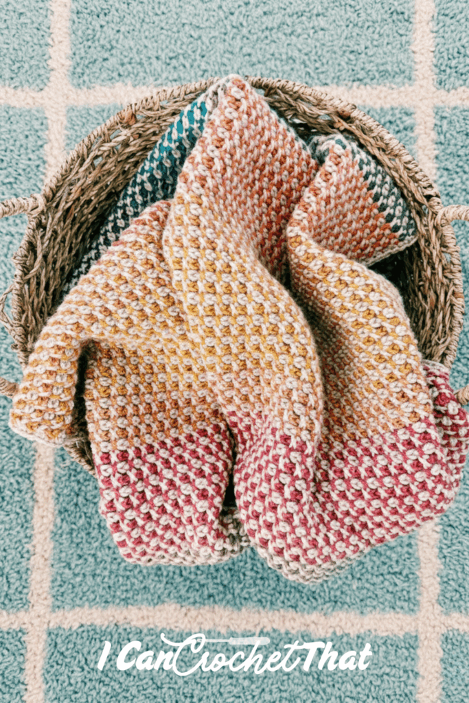 A multicolored crochet blanket with a honeycomb pattern is draped inside a woven basket on a blue and white checkered rug. The text “I Can Crochet That” is at the bottom.